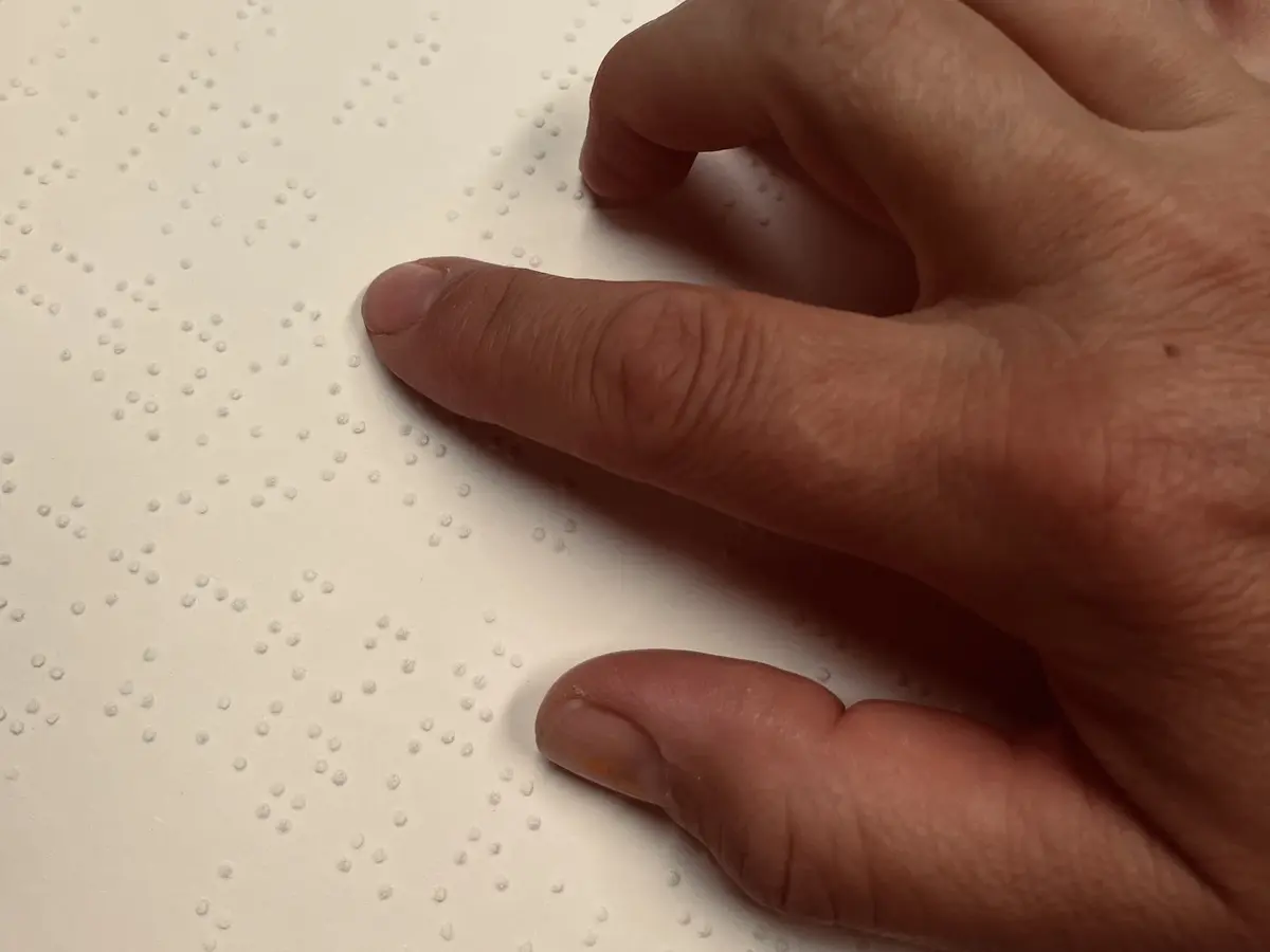 Eine Hand liegt auf einem weißen Blatt mit Braille-Schrift. Der Zeigefinger ist ausgestreckt und lesend auf der Schrift. 