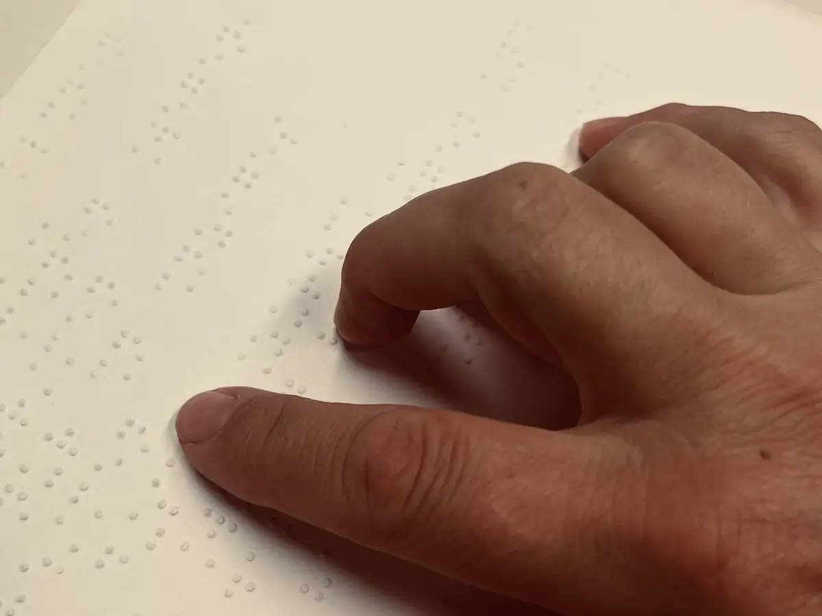 Eine Hand liegt auf einem weißen Blatt mit Braille-Schrift. Der Zeigefinger ist ausgestreckt und lesend auf der Schrift. 